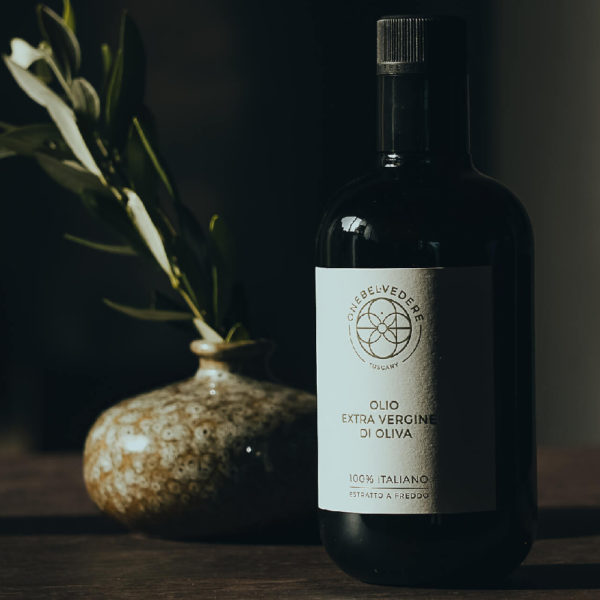 A bottle of Olio, а olive oil from One Belvedere, standing in the shadow on the table with the plant behind it.
