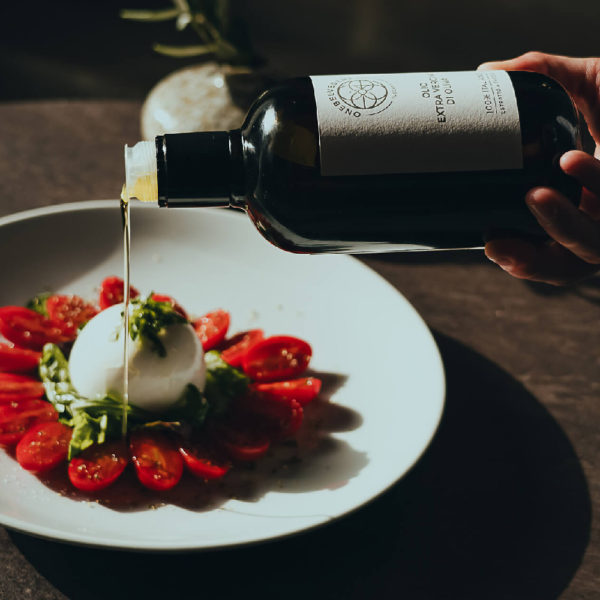 A man's hand holding a bottle of Olio, а olive oil from One Belvedere, and placing it on a food.