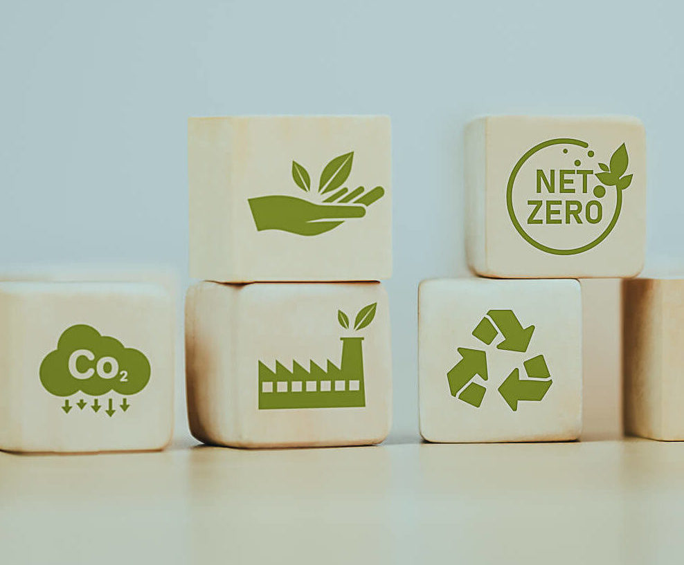 A collection of wooden blocks with various green symbols related to sustainability and environmental protection. These symbols include a hand holding a leaf, the words "NET ZERO" in a circle, a cloud with arrows pointing downward, a factory, and a recycling symbol.