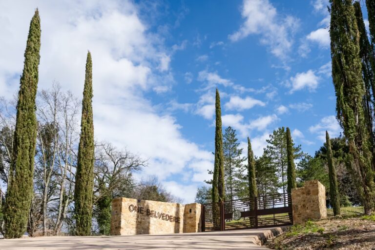 Entrance to One Belvedere Tuscany framed by tall cypress trees beneath a bright Tuscan sky.